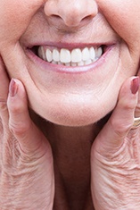 Nose-to-neck view of woman with white hair holding both hands to sides of her face while smiling